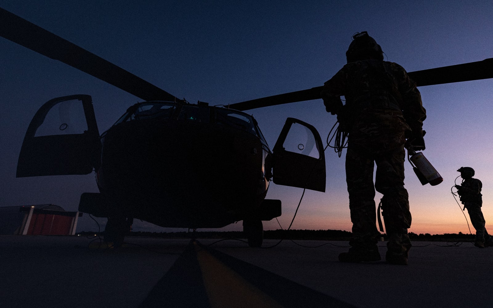U.S. Army National Guard aircrew, seen from behind, silhouetted beside a helicopter at dusk.